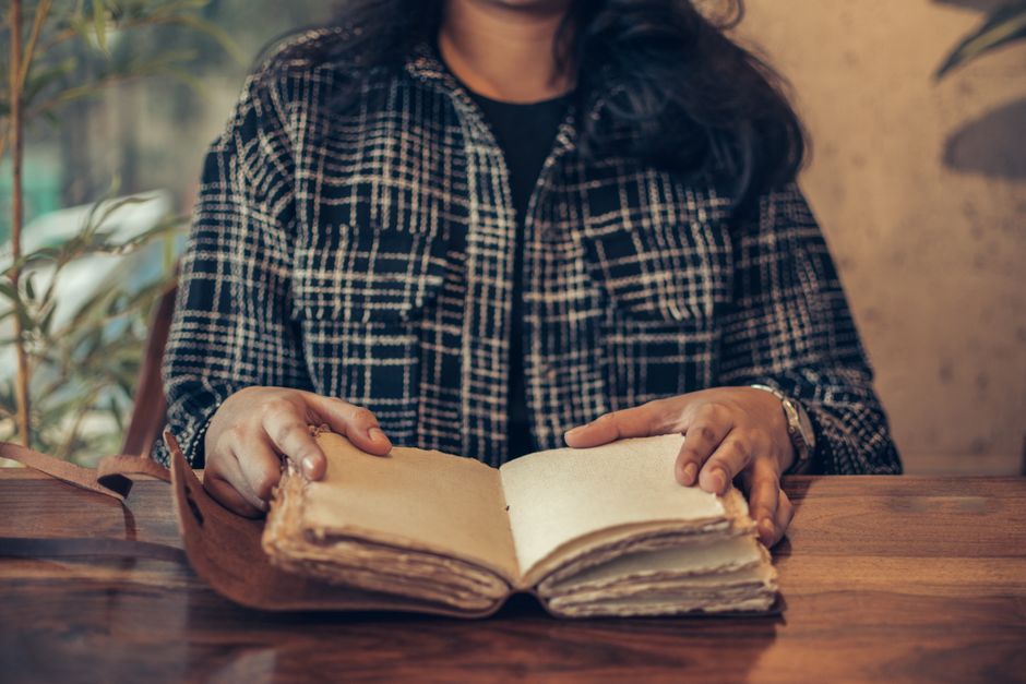 A woman in a plaid shirt reading an antique book at a wooden table.