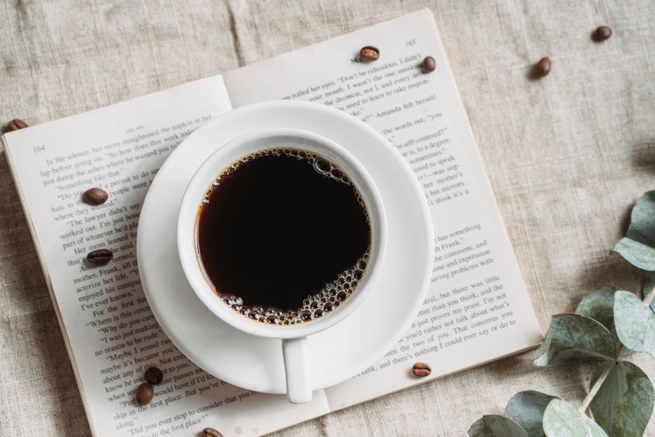 A relaxing setup featuring a cup of black coffee on an open book with scattered coffee beans.
